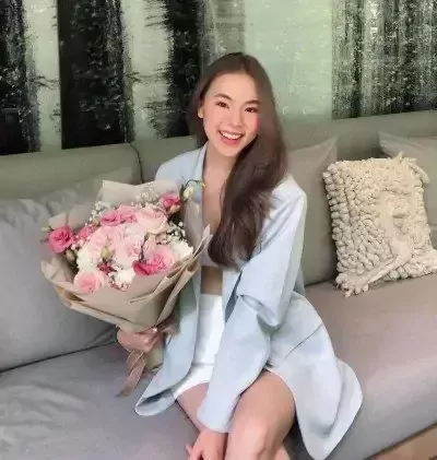 Smiling woman in a light blue blazer holds a bouquet of pink roses and white flowers, sitting on a grey couch.