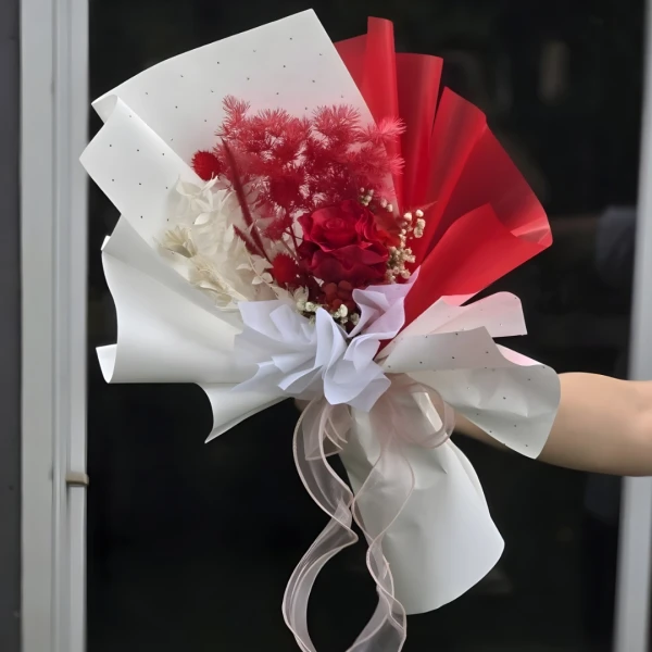 A bouquet of red dried flowers with white limonium, wrapped in white and red paper with a ribbon.