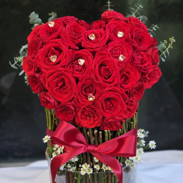 Heart-shaped arrangement of red roses with heart crystals in an acrylic cylinder with a red ribbon.