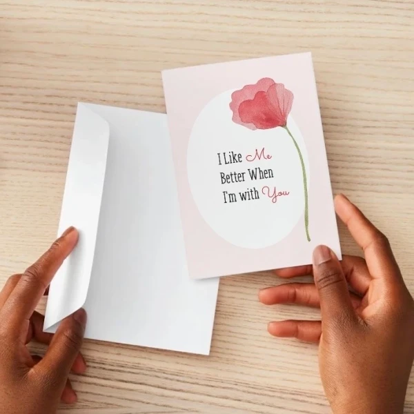 Hands hold a pink card with a red poppy and "I Like Me Better When I'm With You" text next to a white envelope.