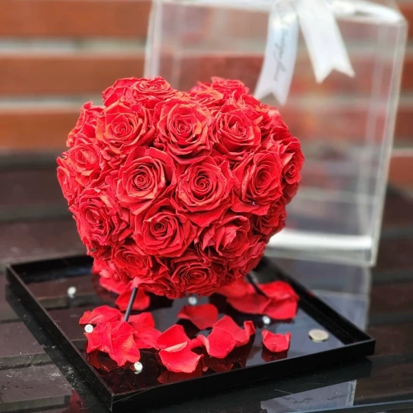 Heart-shaped dome of preserved red roses with scattered petals on a black tray.