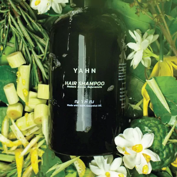 Dark green bottle of YAHN Hair Shampoo surrounded by fresh herbs and white flowers.