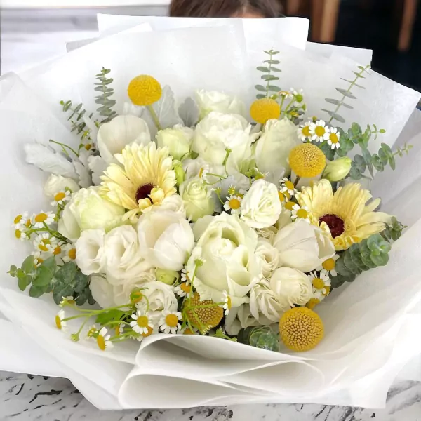 Cream gerberas, white roses, yellow craspedia, daisies, and lisianthus in a white paper bouquet.