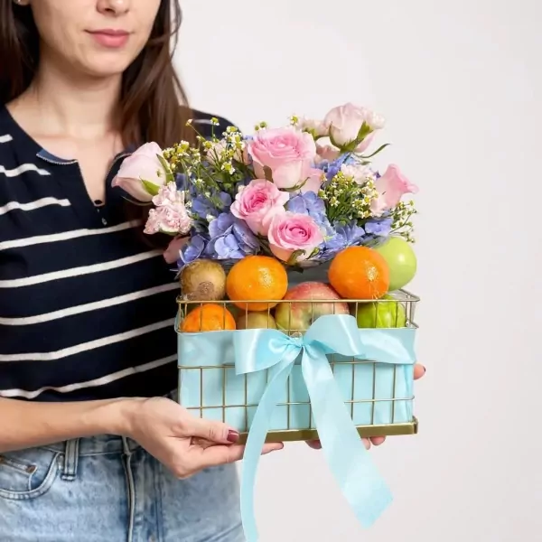 A woman holds a light blue gift basket filled with fruits like oranges and apples, topped with pastel pink roses and blue hydrangeas.