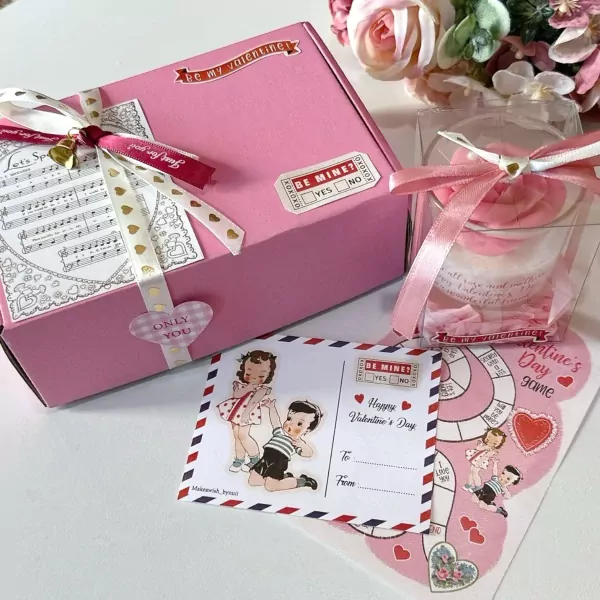 Pink gift box with ribbon, rose candle in clear box, and Valentine's Day cards and stickers.
