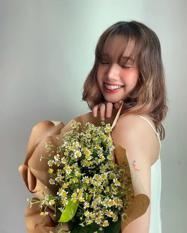 Woman smiling, holding a bouquet of white daisies wrapped in brown paper.