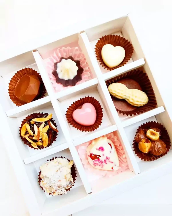 A white box filled with nine handcrafted chocolates of various shapes and toppings, including nuts and fruit.