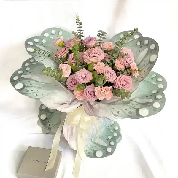 Green butterfly bouquet with pink roses and carnations, tied with a cream ribbon, next to a Charles & Keith gift box.