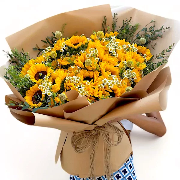 A large bouquet of sunflowers, accented with small white flowers and greenery, wrapped in brown paper.