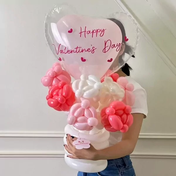A person holds a balloon arrangement with a large pink heart balloon saying "Happy Valentine's Day" above balloon roses in a white vase.