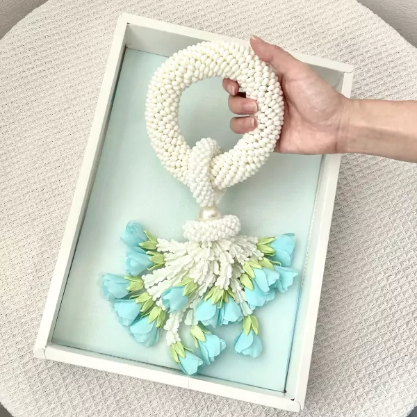 A white beaded jasmine malai with light blue flowers is held in a hand above a white frame.