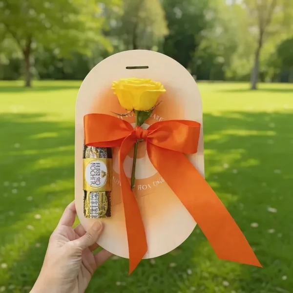 A hand holds a graduation gift of a yellow rose and two Ferrero Rocher chocolates tied with an orange ribbon.