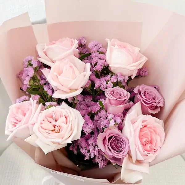 A pastel bouquet of pink and purple roses with purple statice, wrapped in soft pink paper.