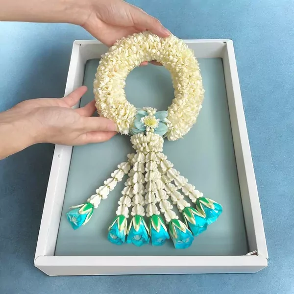 Jasmine malai garland with blue flower accents in a white box, held by hands.