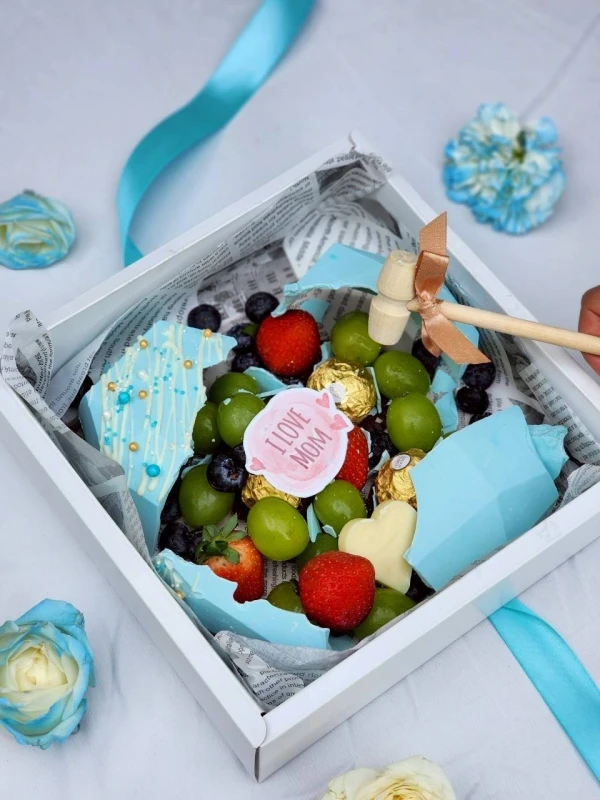 Blue smash heart cake with fruits, chocolates, and a small hammer, presented in a white box.