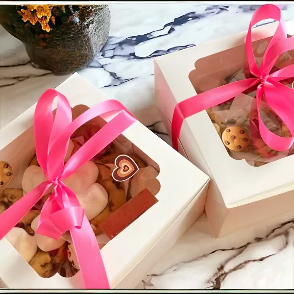 Two white gift boxes filled with chocolate chip cookies, tied with pink ribbons, on a marble surface.