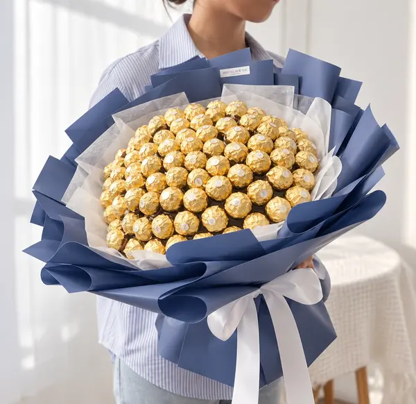 A person holds a large bouquet made of 100 Ferrero Rocher chocolates wrapped in navy blue and white paper.