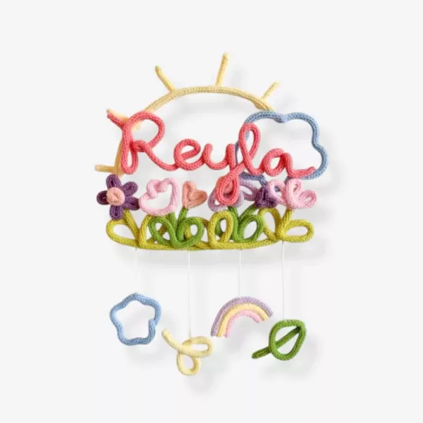 Knitted wall hanging with "Reyla" in pink and coral, sun, cloud, flowers, and hanging rainbow, star, and leaf.