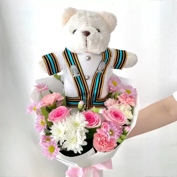 White teddy bear in a Chula graduation gown surrounded by pink and white flowers.