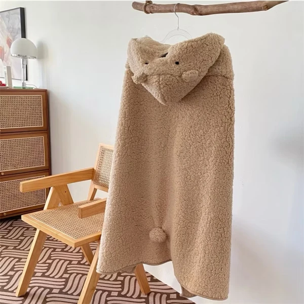 A beige teddy hoodie blanket with bear ears and a pom-pom hangs on a rod above a wooden chair.
