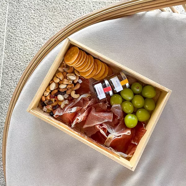 Charcuterie box with Serrano ham, crackers, nuts, dried fruit, grapes, and jam jars.