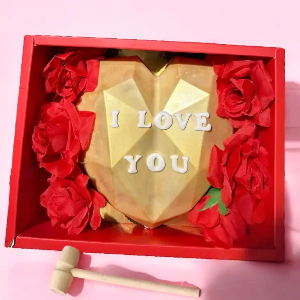 Golden heart-shaped chocolate piñata with "I LOVE YOU" spelled out, surrounded by red roses in a red box with a wooden hammer.