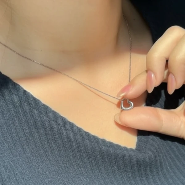 Hand with nails touches a tiny silver heart pendant necklace on a woman's neck.