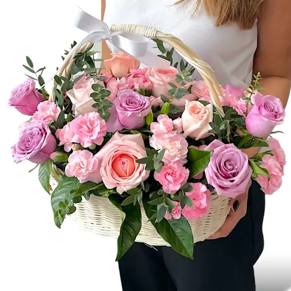 A white woven basket brimming with pink and purple roses, carnations, and greenery, tied with a white ribbon.