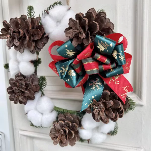 Christmas wreath with pine cones, cotton, and festive red and green bows on a white door.