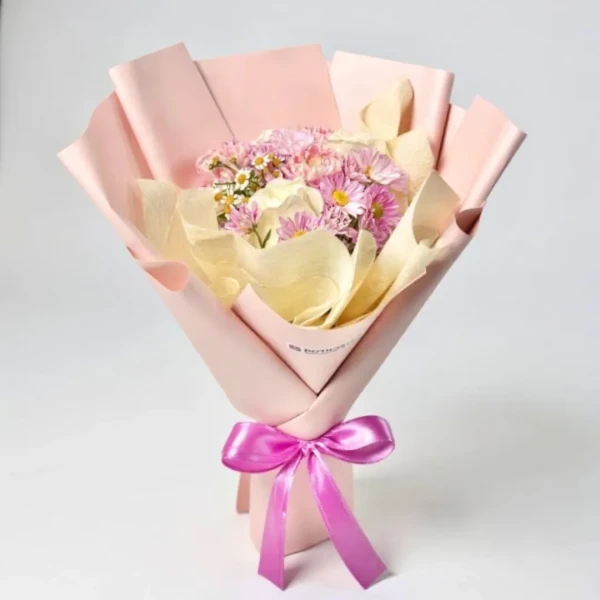 Bouquet of pink daisies, white roses, and chamomile in blush paper, tied with a pink bow.