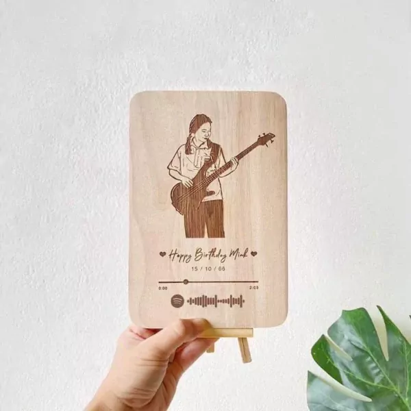 Wooden plaque with laser-engraved portrait of person playing guitar, song title, date, and scannable music code.