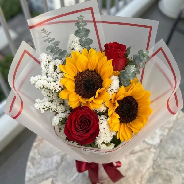 Bouquet of sunflowers and red roses with white fillers and eucalyptus, wrapped in translucent paper and tied with a red ribbon.