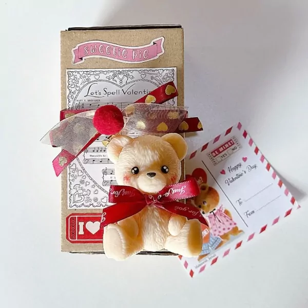 Teddy bear candle with red ribbon, set in a box with Valentine's Day decor and a postcard.