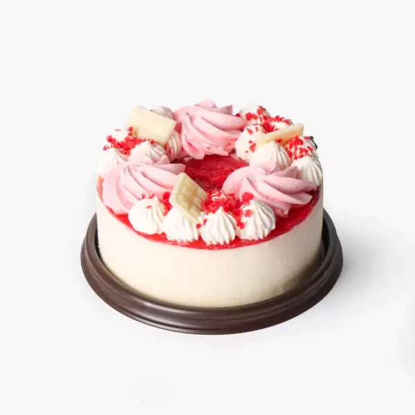 Round strawberry yogurt cake topped with pink and white frosting, white chocolate, and red sprinkles on a dark base.