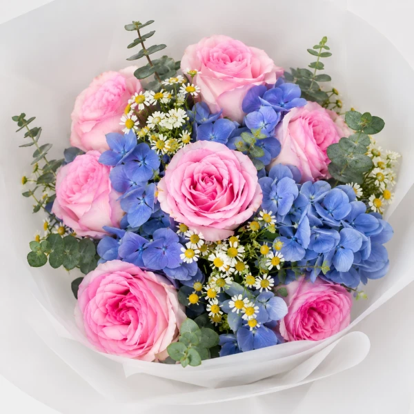 Pink roses, blue hydrangeas, and daisies in white paper wrap, tied with a dark blue ribbon.