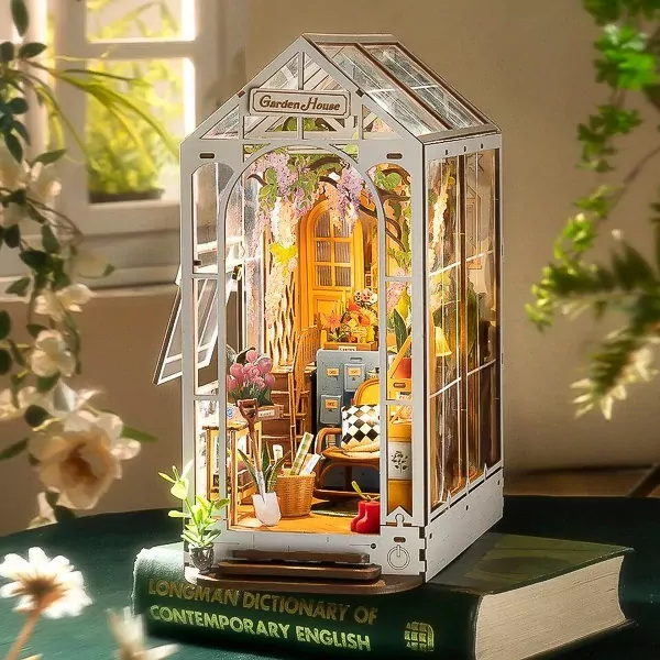 A miniature wooden greenhouse book nook with plants, furniture, and flowers, sitting on a book.