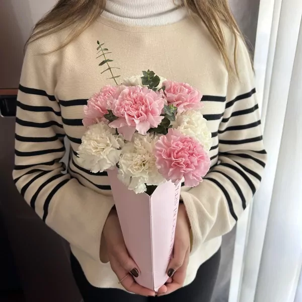 A person in a striped sweater holds a pink and white carnation bouquet in a pink paper vase.