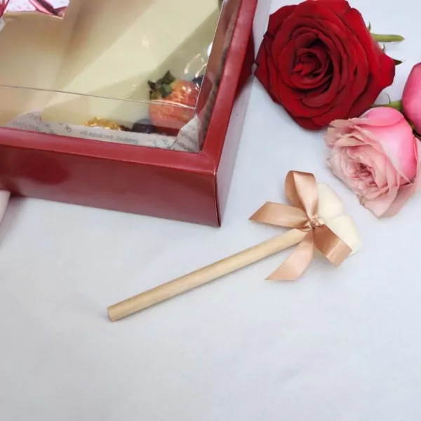 White chocolate heart cake pop with a bow on a wooden stick, next to a red box and roses.