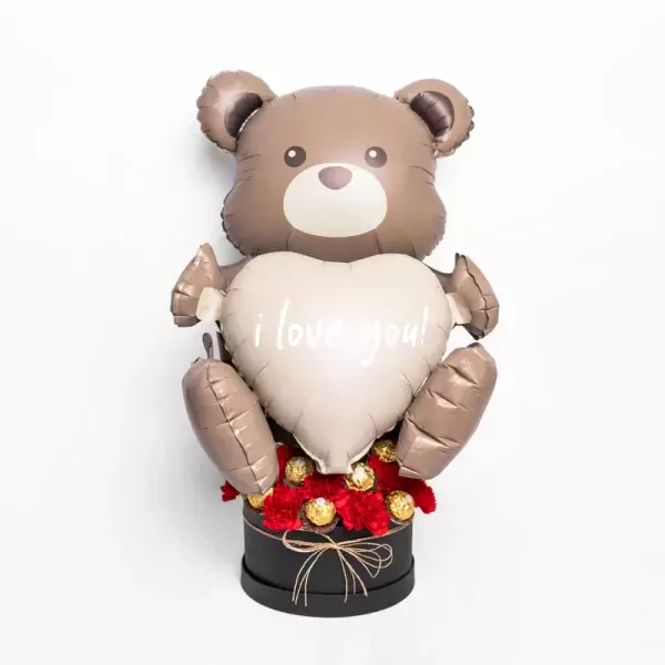 Teddy bear balloon holding a heart with "i love you!" message, in a box with red carnations and chocolates.
