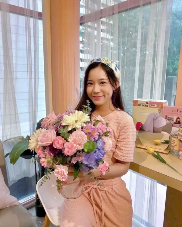 A young woman in peach loungewear holds a pastel flower bouquet with blue hydrangeas, pink roses, and white gerberas.