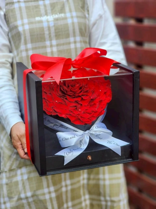A person holds a black box with a red preserved rose, red bow, and Ferrero Rocher chocolates.