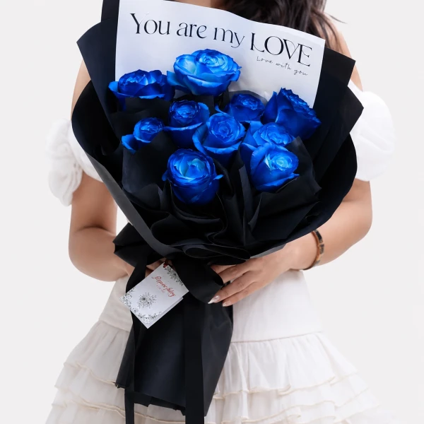 A bouquet of vibrant blue roses wrapped in black paper, held by a person in a white dress.