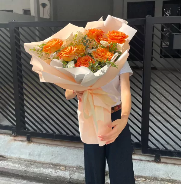 A bouquet of orange roses, alstroemerias, and daisies wrapped in light pink paper.