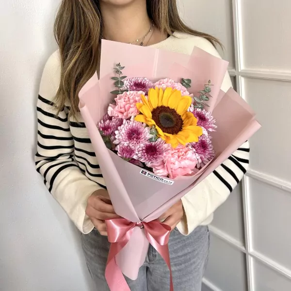 A woman holds a bouquet featuring a bright yellow sunflower, pink carnations, and purple chrysanthemums.