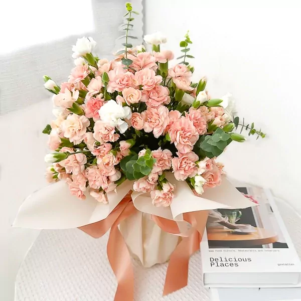 Pink carnation and white flower bouquet in a vase, tied with a ribbon, next to a book.