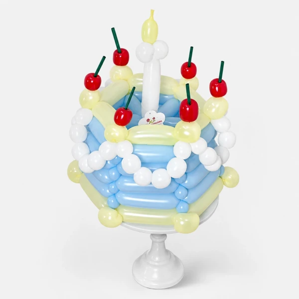 A blue and yellow balloon cake with white balloon accents, red cherries, green stems, and yellow candle balloons on a white stand.