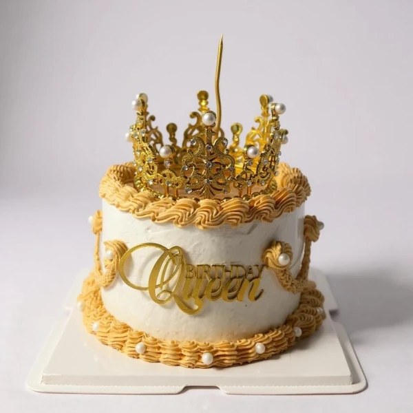 Gold crown cake with "Queen" topper, white frosting, and gold piping.