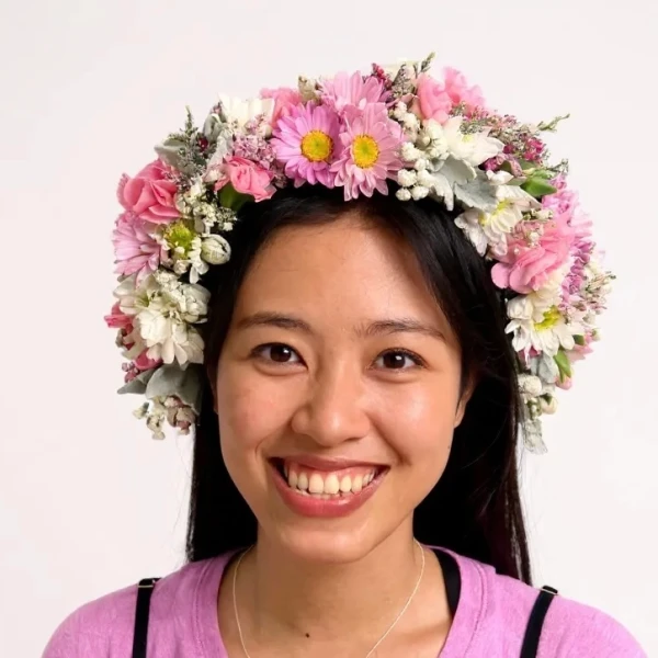 A smiling young woman wears a large, colorful fresh flower crown with pink, white, and yellow blooms.