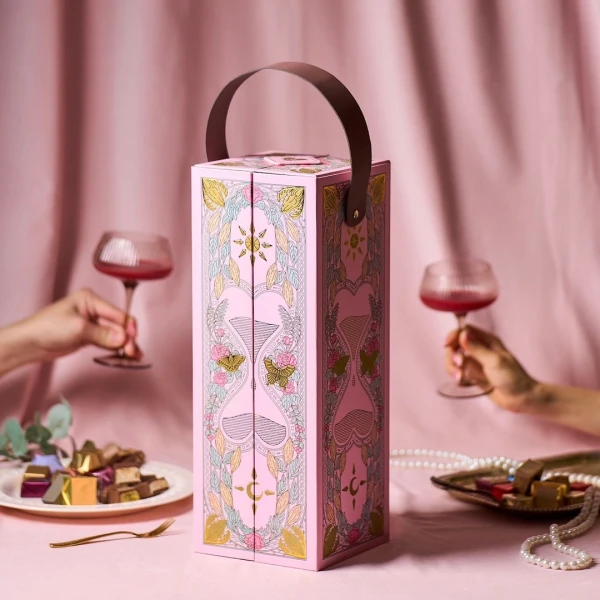 Pink chocolate box with romantic floral and hourglass designs, holding chocolates, with hands holding wine glasses.