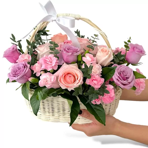 White wicker basket overflowing with pink, purple, and blush roses, carnations, and greenery.